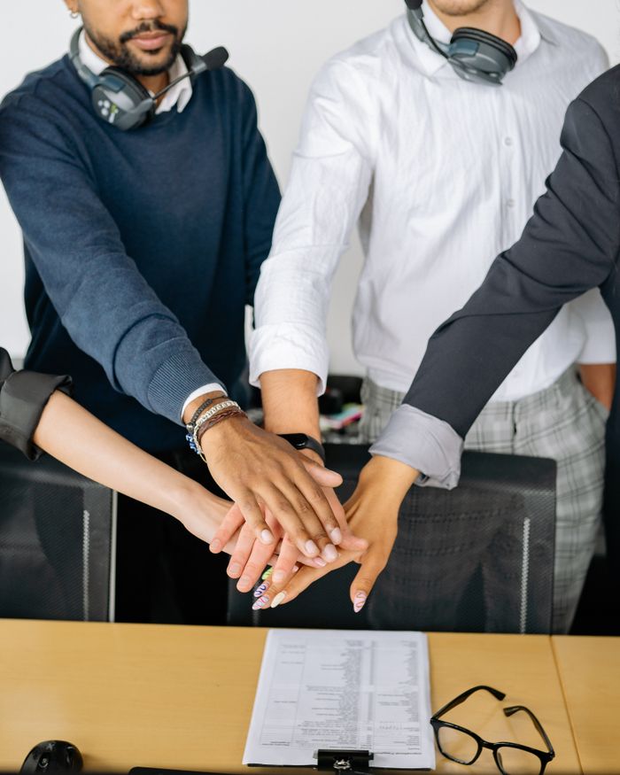 our-story-01 Diverse colleagues joining hands on conference table, symbolizing teamwork and unity.